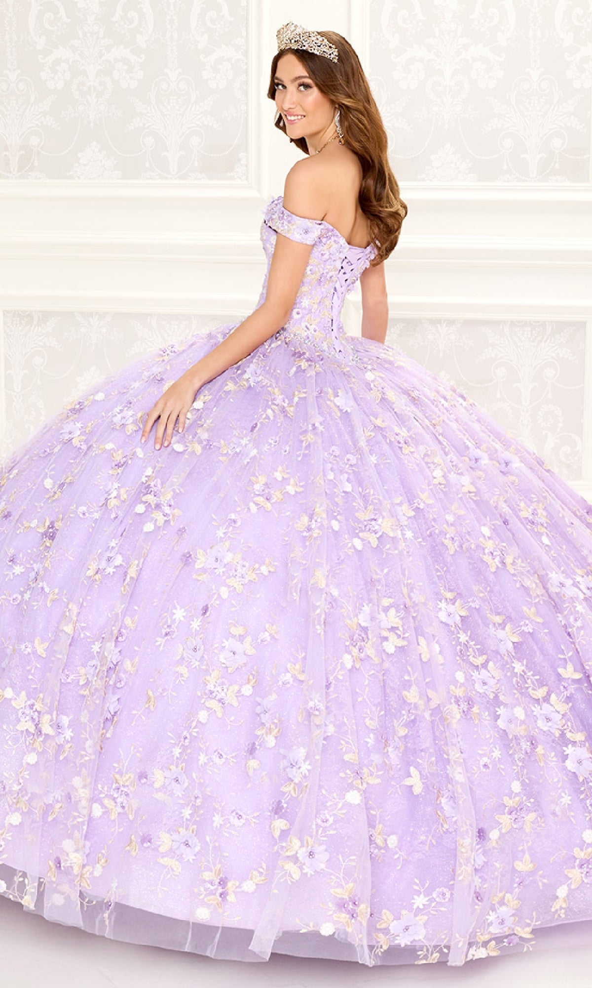 A woman wearing the Quinceanera Dress PR30084 By Princesa, featuring floral embellishments and a corset back, stands in a decorated white room with a tiara, facing slightly away from the camera.